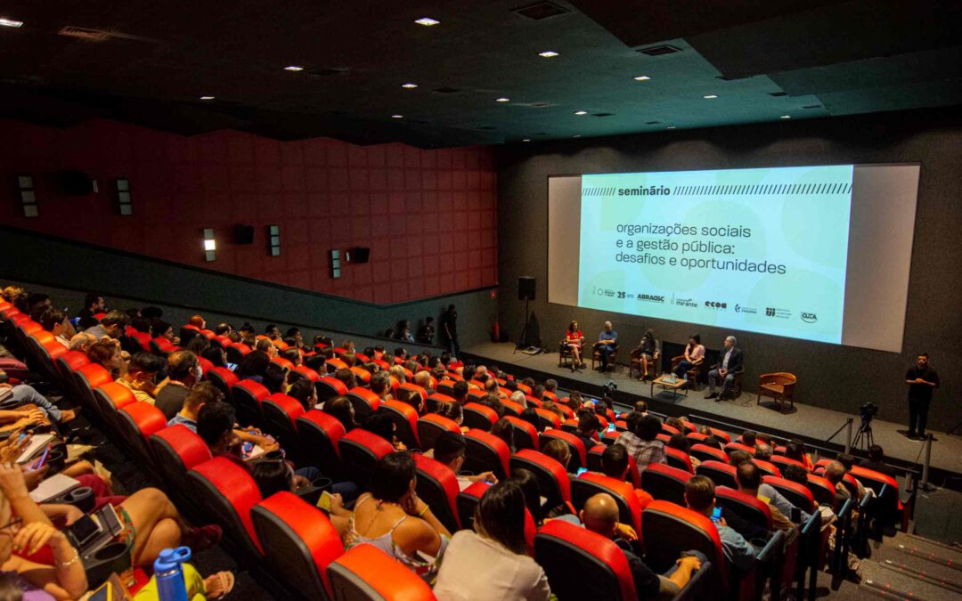 Em um cinema de mais de 100 lugares, lotado, o público est sentado em cadeiras ´vermelhas e à frente os palestrantes estão sentados abaixo da tela de exibição das apresentações.
