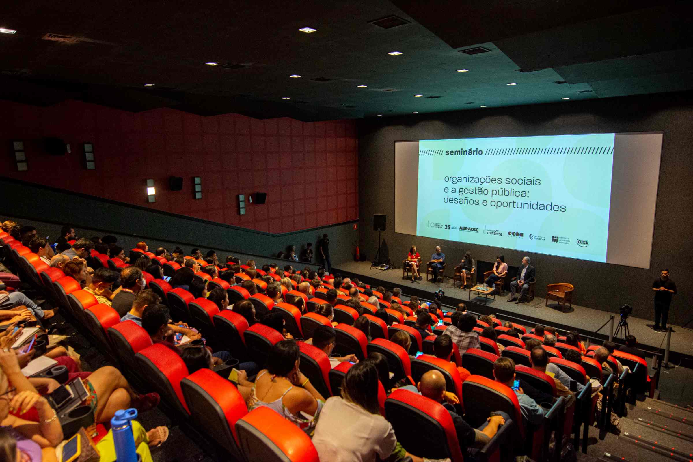 Em um cinema de mais de 100 lugares, lotado, o público est sentado em cadeiras ´vermelhas e à frente os palestrantes estão sentados abaixo da tela de exibição das apresentações.