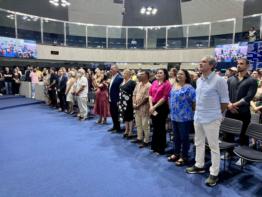 Várias pessoas na Assembleia Legislativa do Estado do Ceará, roupas, estilos e etnias diversos, em posição de respeito à execução do Hino Nacional.