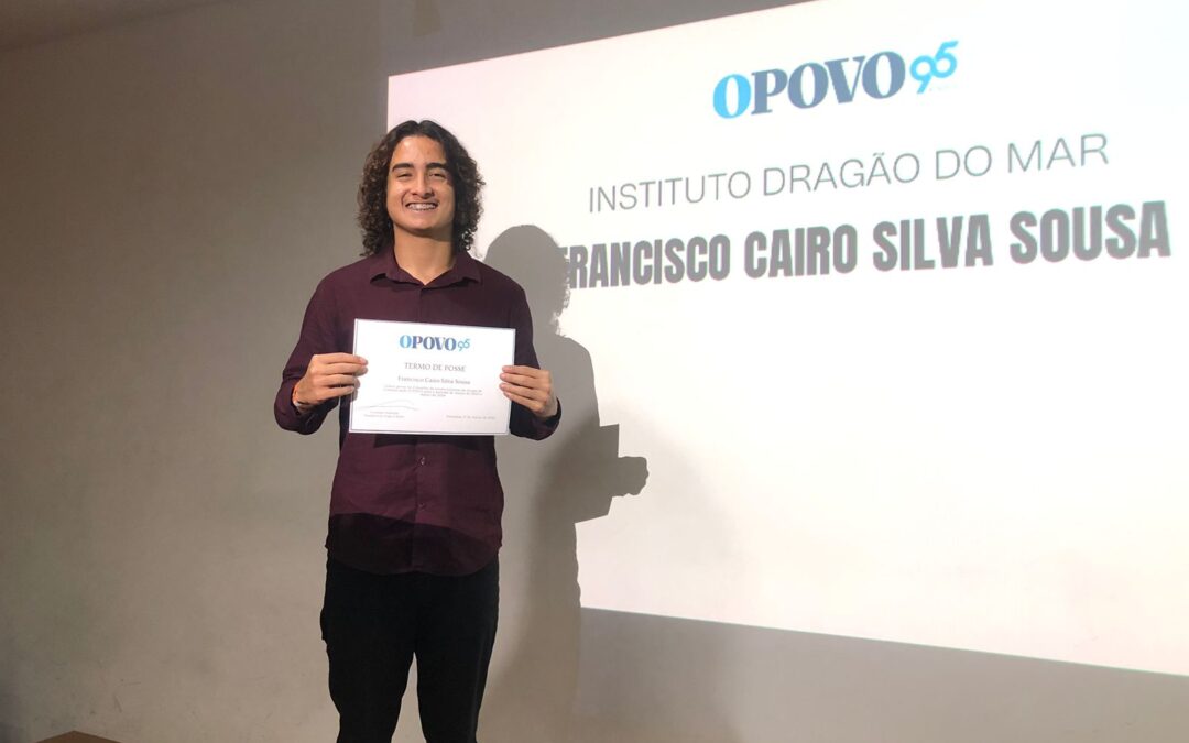 Um jovem de camisa vinho e calça preta, cabelo cacheado na altura dos ombros, sorri para a câmera segurando um certificado em que se lê O POVO. Ao fundo, a logo do jornal O POVO em azul e, abaixo: Instituto Dragão do Mar e Francisco Cairo Silva Sousa.