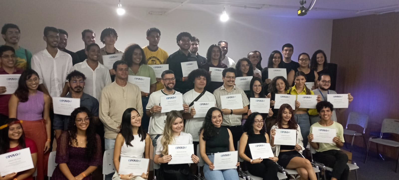 Vário jovens, de etnias diversas, com roupas de estilos e cores diferentes seguram nas mãos um certificado em que é legível "O POVO".