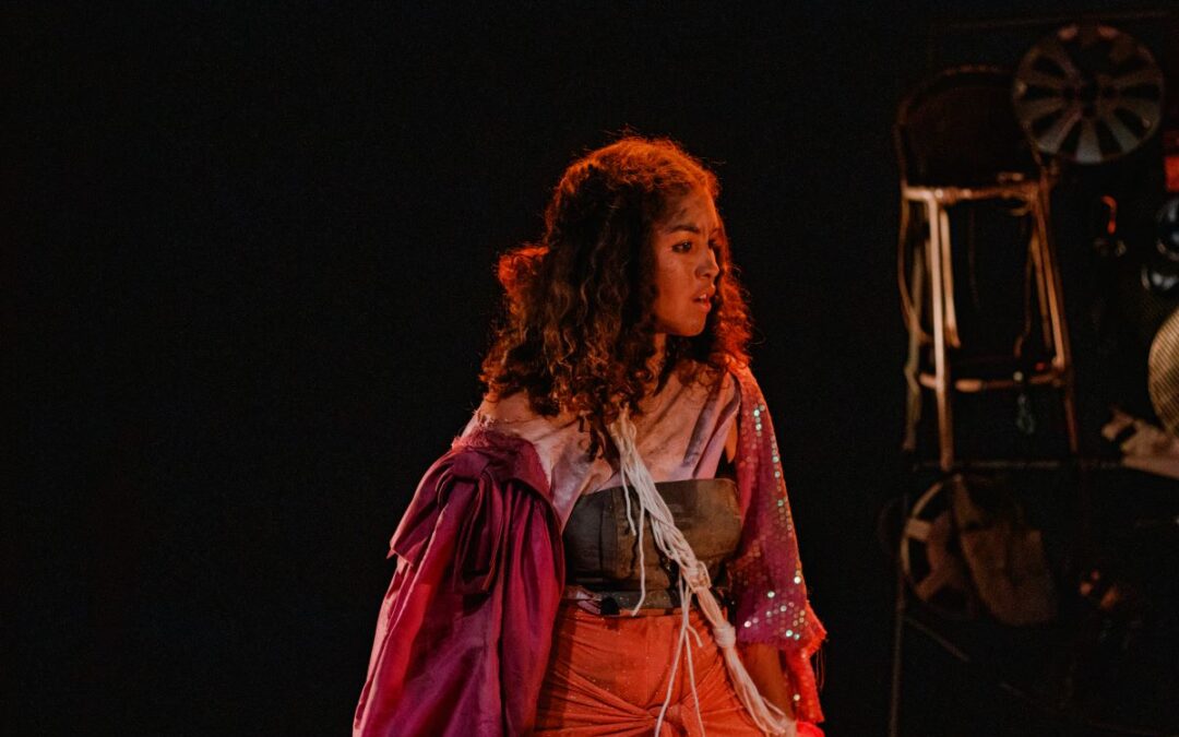 Uma jovem de cabelos cacheados com roupa de várias cores está no centro do palco e da foto, olhando para o lado