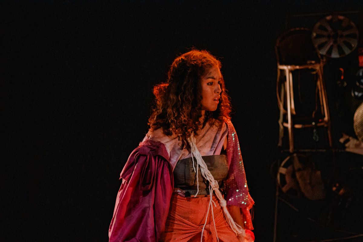 Uma jovem de cabelos cacheados com roupa de várias cores está no centro do palco e da foto, olhando para o lado