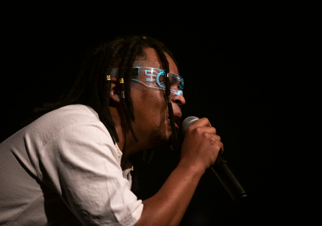 Foto de homem negro cantando. Ele usa óculos transparente com detalhes azuis.