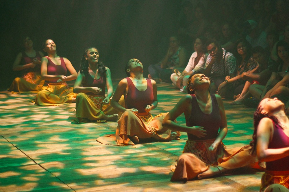 Um grupo de mulheres sentadas no chão com maiôs vermelhos e saias amarelas. O público assiste ao redor. 