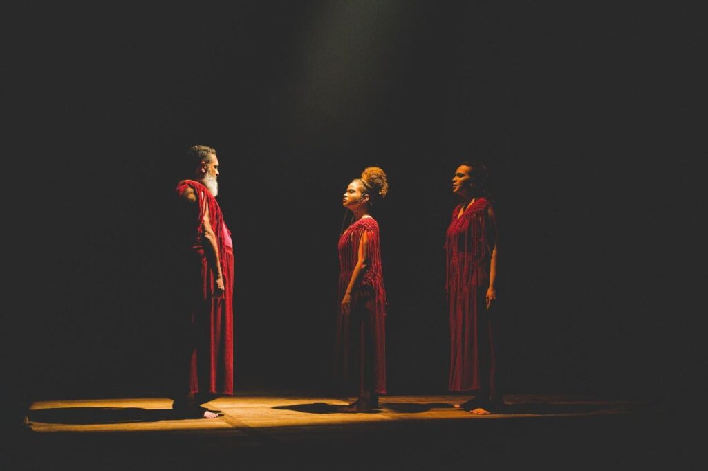 Três pessoas de vermelho num palco escurecido conversam.