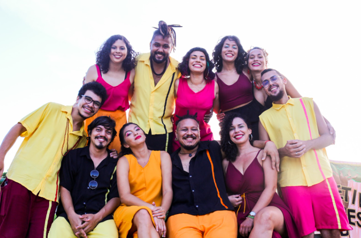 Foto do grupo Raízes do Girô. São 11 pessoas. Todas usam mesma paleta de cores, incluindo rosa, amarelo, preto e vinho. Todos estão sorrindo.