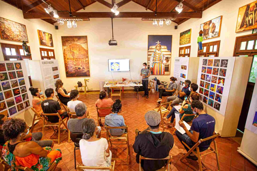 Uma sala de museu, com diversos quadros, diversas pessoas assistem um homem falando sobre um tema.