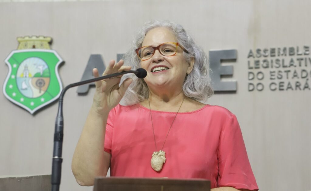 Rachel Gadelha, uma mulher cis, cabelos brancos, óculos de aro tartaruga, colar em formato de coração e blusa rosa fala ao microfone fixo de um pequeno  púlpito.