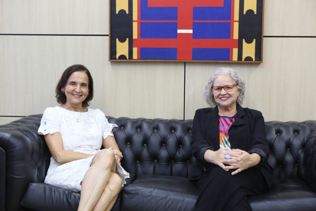 uma mulher branca, de vestido branco, cabelos curtos castanhos senta-se ao lado de outra mulher branca, cabelos curtos brancos, blazer preto, calça preta e blusa estampada, em sofá preto  com a metade de um quadro em vermelho, preto azul e laranja atrás delas. 