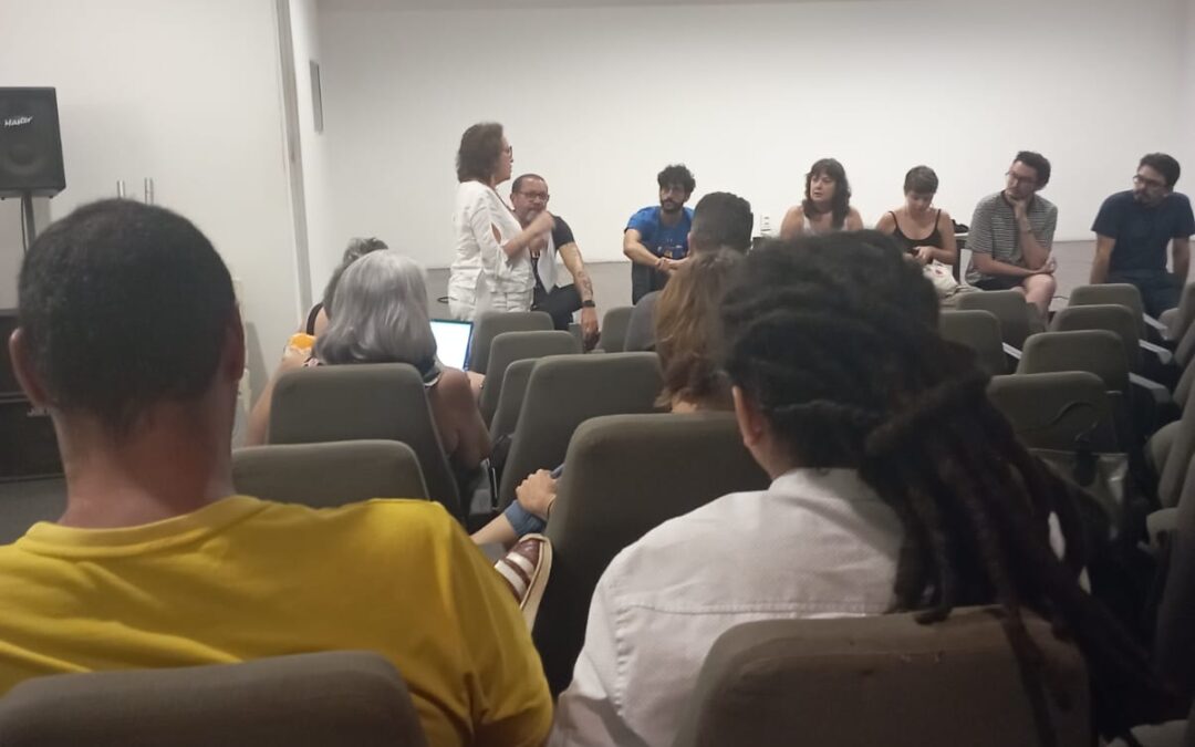Diversas pessoas em um auditório assistem à fala de uma mulher vestida de branco que segura o microfone está quase de costas para a plateia. Sentados e sentadas no palco, algumas pessoas asssitem e dialogam com ela. Os presentes vestem roupas de cores variadas e pertencem a diferentes raças.
