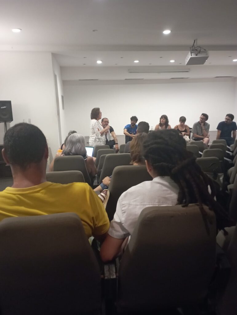 Diversas pessoas em um auditório assistem à fala de uma mulher vestida de branco que  segura o microfone  está quase de costas para a plateia. Sentados e sentadas no palco, algumas pessoas asssitem e dialogam com ela. Os presentes vestem roupas de cores variadas e pertencem a diferentes raças. 