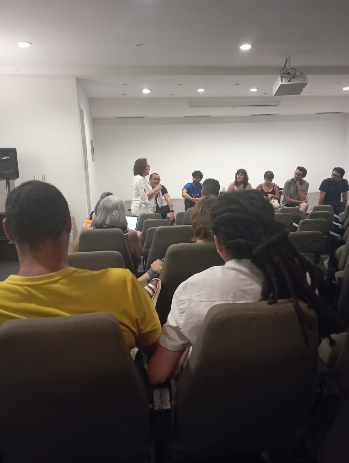 Diversas pessoas em um auditório assistem à fala de uma mulher vestida de branco que segura o microfone está quase de costas para a plateia. Sentados e sentadas no palco, algumas pessoas asssitem e dialogam com ela. Os presentes vestem roupas de cores variadas e pertencem a diferentes raças.