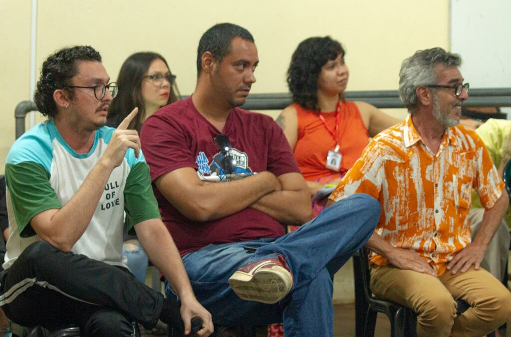 Sentados em cadeiras pretas, de plástico, comuma parede bege ao fundo, diversas pessoas, de diferentes raças, roupas de diferentes cores, escutam.