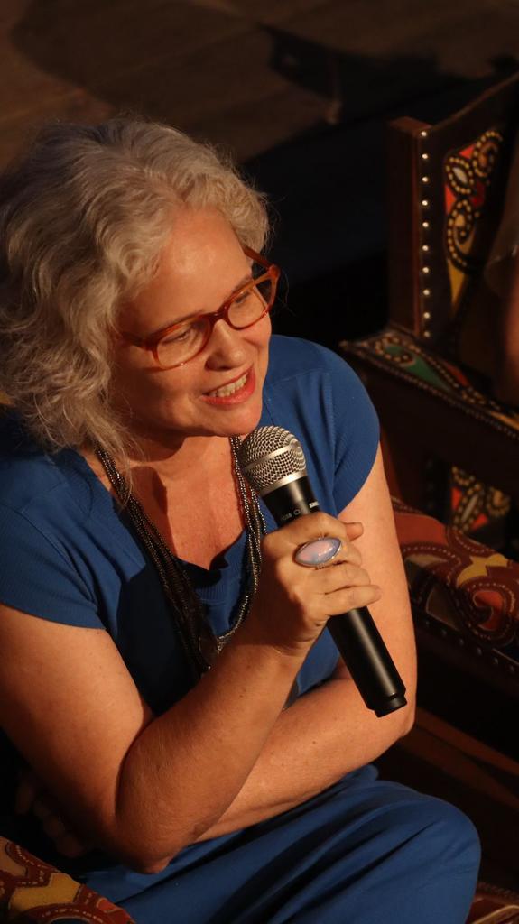 Uma mulher branca, cabelos brancos, de óculos, roupa azul fala ao microfone.
