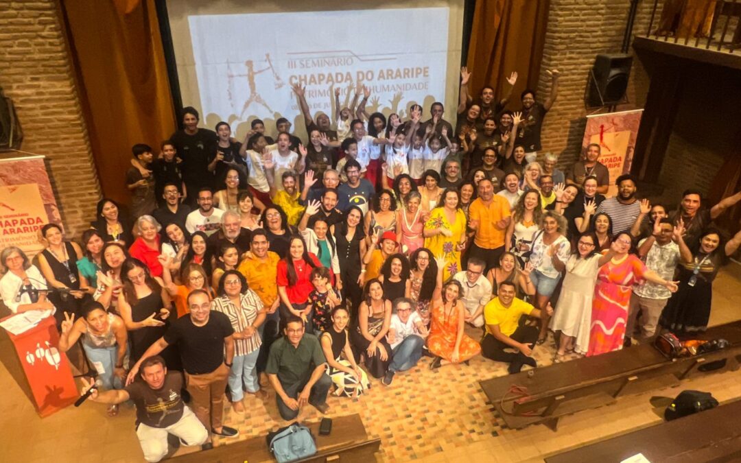 Em um placo baixo, pessoas de diversas raças e roupas em diferentes cores, Está reunida. Algumas têm as mãos levantadas.