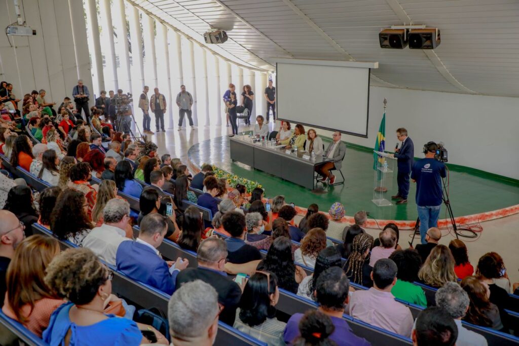 Em um auditório lotado quatro pessoas estão à frente da plateia em uma mesa e um homem de terno discursa ao lado em um púlpito transparente   