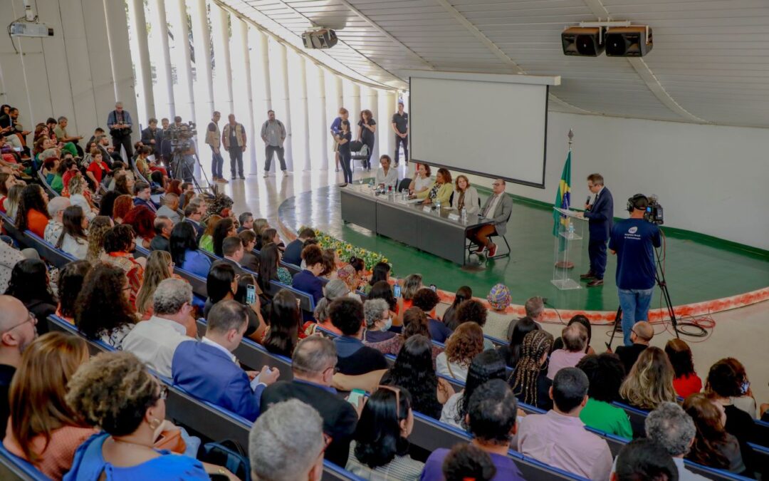 Em um auditório lotado quatro pessoas estão à frente da plateia em uma mesa e um homem de terno discursa ao lado em um púlpito transparente