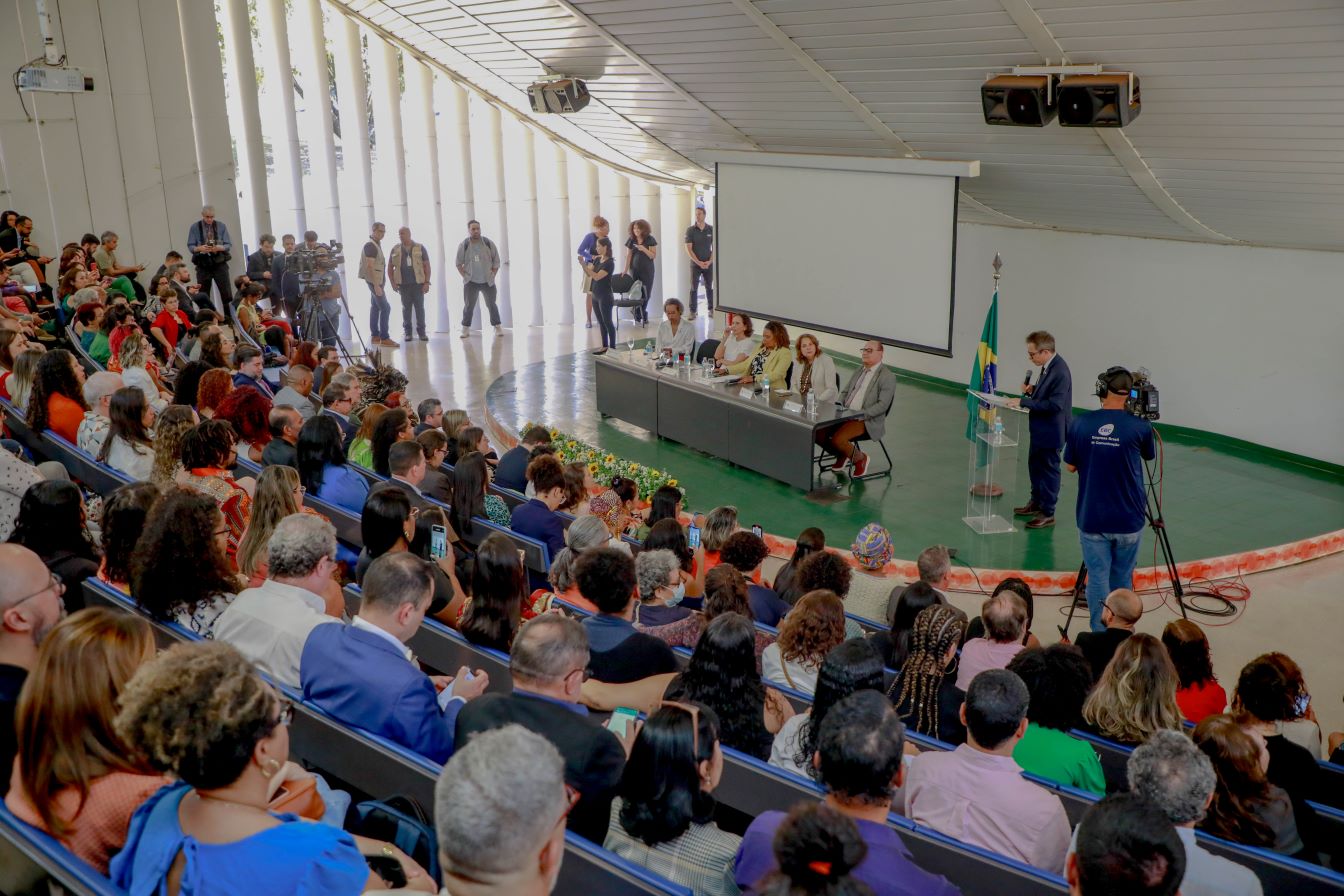 Em um auditório lotado quatro pessoas estão à frente da plateia em uma mesa e um homem de terno discursa ao lado em um púlpito transparente