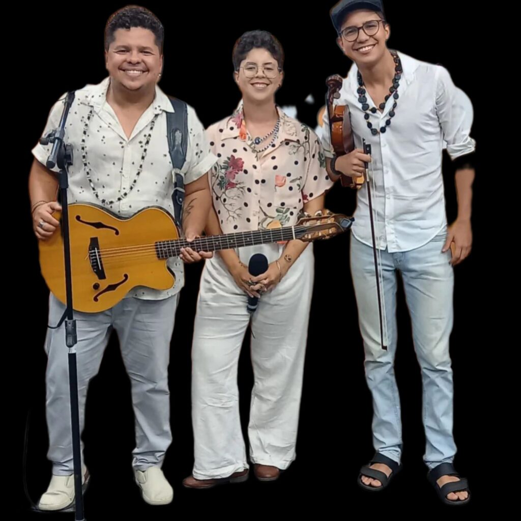 Um trio com instrumentos musicais diversos e roupas de cores variadas posa para a foto, de corpo inteiro.