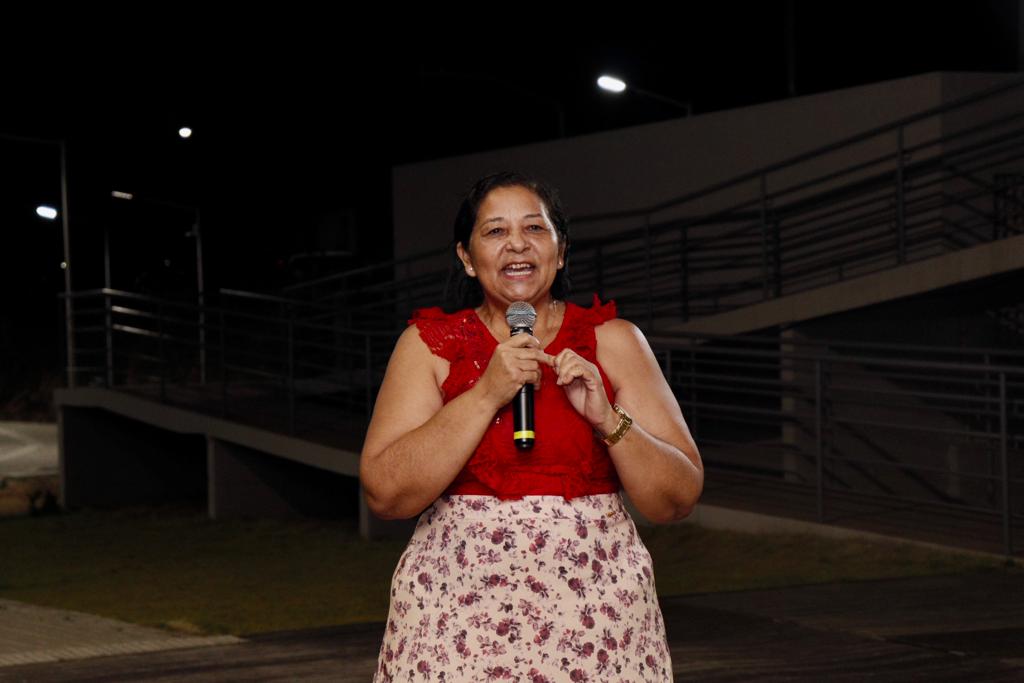 uma mulher de cabelos pretos curtos, camisa vermelha e saia estampada em vermelho fala ao microfone