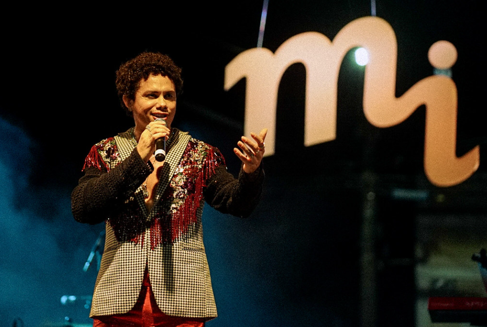 Foto de Silvero Pereira. Ele usa terno estampado e calça vermelha. Está cantando com microfone próximo a boca. Atrás dele, está um letreiro grande com a sigla do festival "MI".