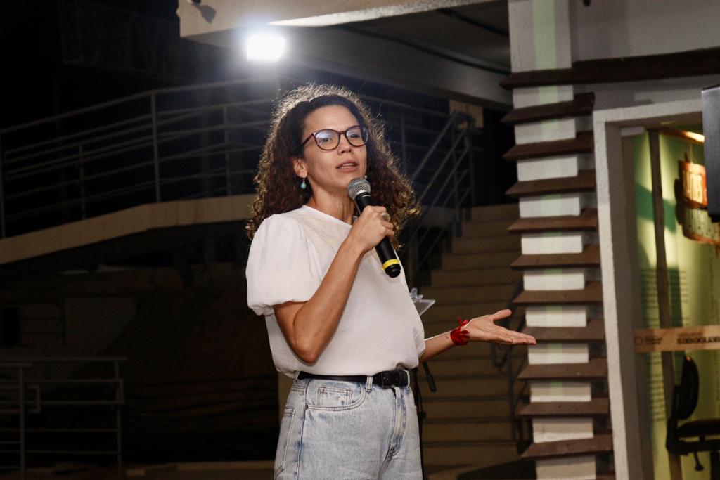 mulher branca, de óculos, cabelos longos, castanhos e cacheados, de blusa branca e calça jeans, fala ao microfone