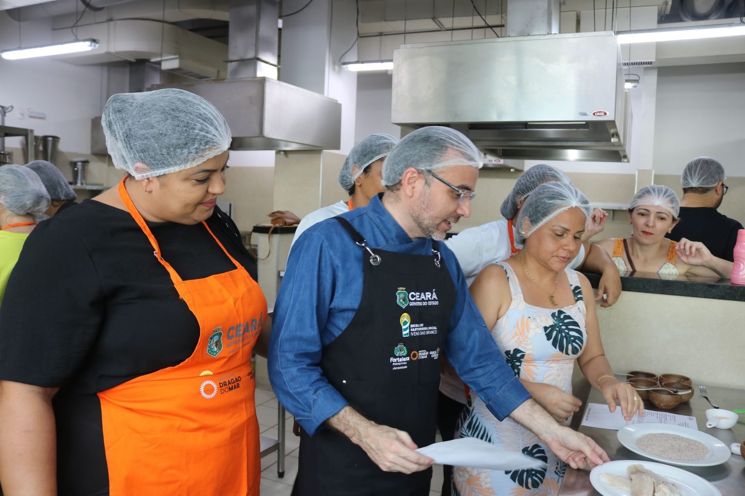 Em uma cozinha industrial, um homem branco, de touca e avental azul-marinho ministra uma aula de gastronomia para diversos alunos e alunas.