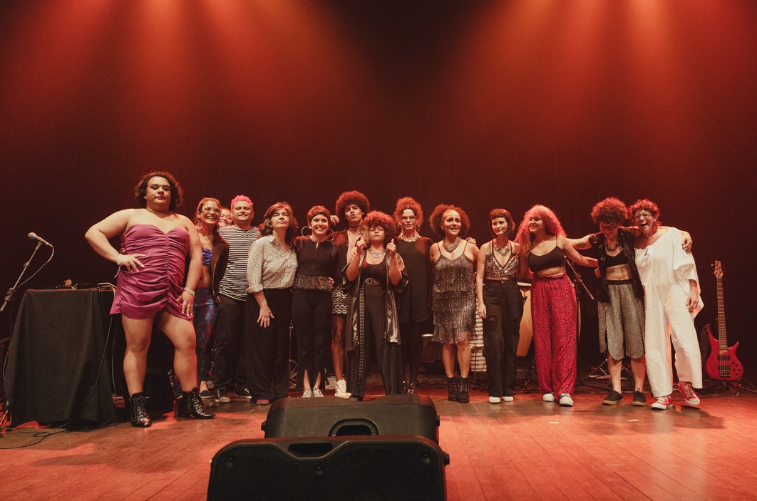 Sob uma luz avermelhada, pessoas de diversas raças, com roupas de cores variadas, posam para a foto no palco. Algumas estão sérias, outras sorriem.