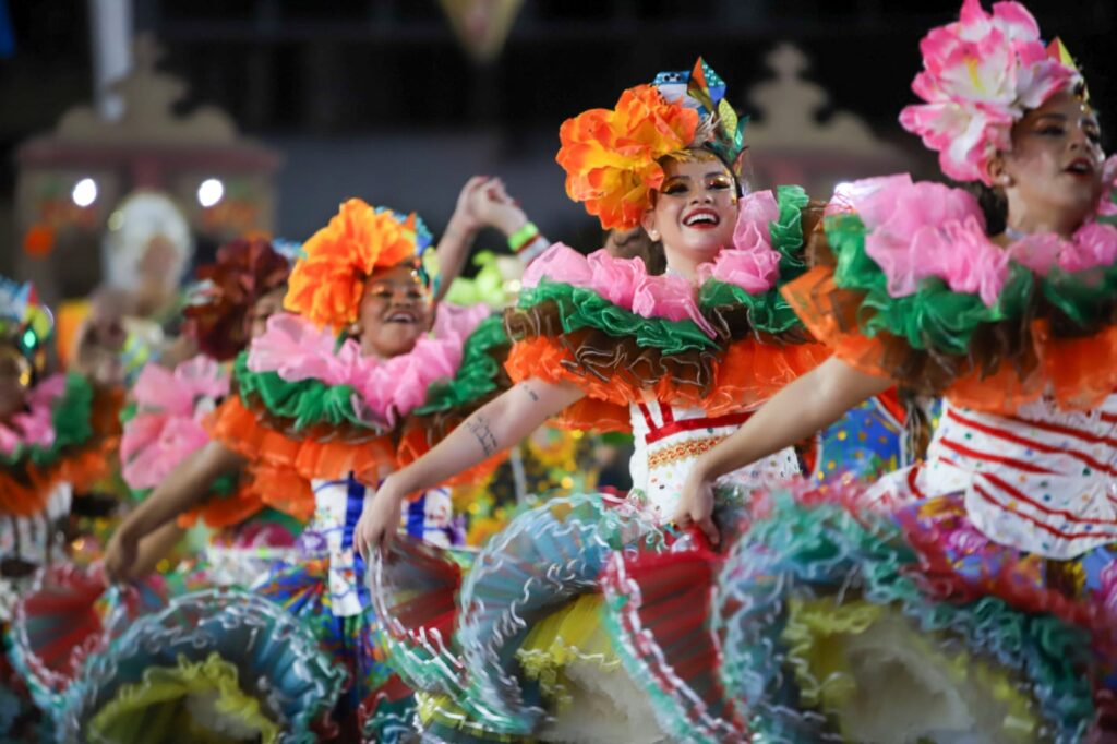 Foto de grupo de quadrilha. Usam vestidos típicos coloridos. Apenas mulheres aparecem nas fotos. Todas sorriem e aparentam cantar. 