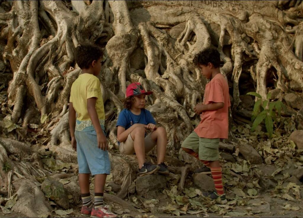 Três garotos (um branco e dois negros),de roupas em cores variadas, conversam em círculo próximos a uma árvore frondosa. Um deles(de camisa azul, short cáqui e boné vermelho) está sentado na raiz da árvore. Os dois outros estão virados para ele, em pé.