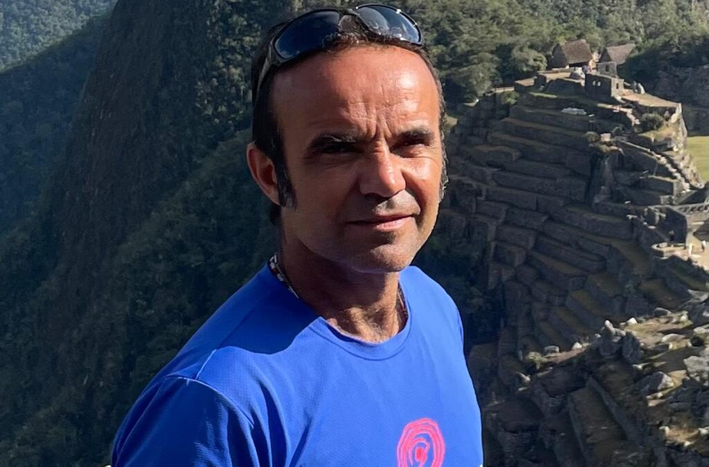 Um homem pardo, de óculos na testa e camiseta azul encara a câmera, em meio à spedras que circundam um penhasco