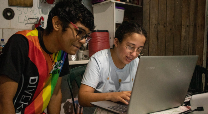 Foto de dois jovens em frente ao computador. Eles parecem estar em casa. No fundo é possível ver uma pia e um armário, o que faz crer que estão na cozinha. Ambos usam oculos e olham atentamente para a tela. 