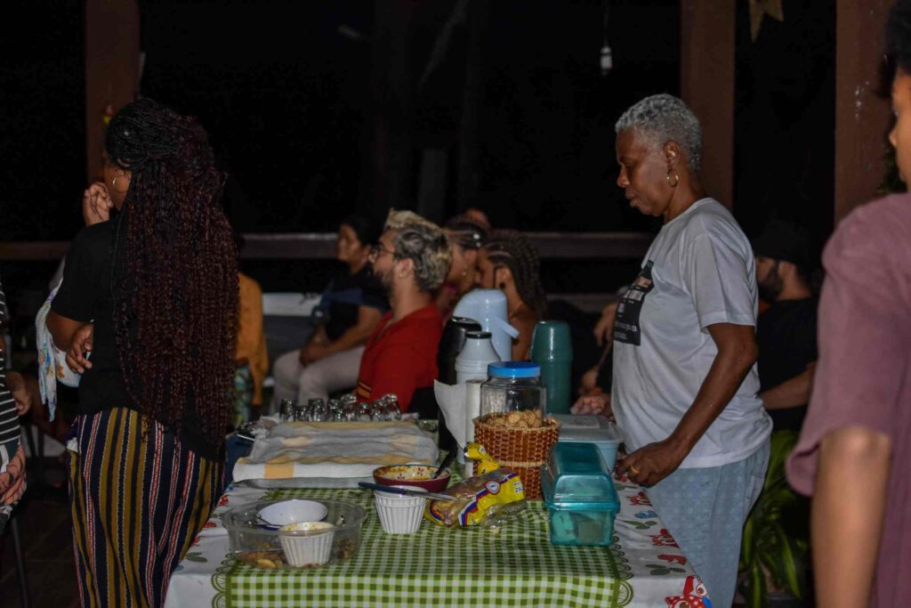 Um grupo de pessoas assistem algo sentadas. Uma mulher negra, de cabelos brancos, curtos, se destaca em frente a uma mesa  de salgados e outros petiscos.