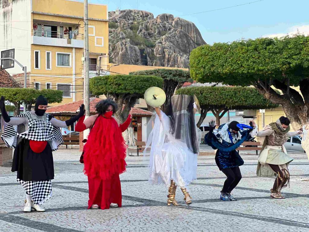 Cinco artistas com roupas e adereços coloridos, de mãos levantadas e cabeças baixas, dançam em uma praça.