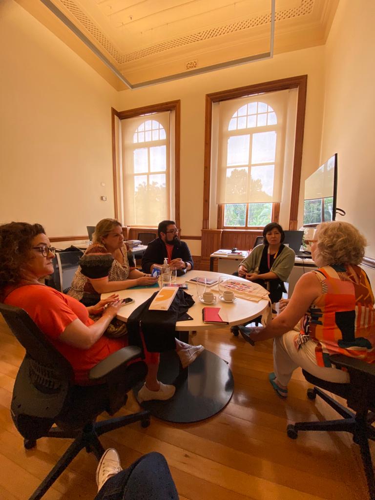 Numa sala, com janelas que se abrem pro sol, em torno de uma mesa redonda, quatro mulheres e um homem  dialogam. 