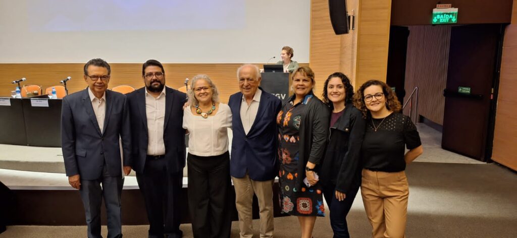 Em frente a um palco baixo, sete pessoas brancas (3 homens e 4 mulheres) com roupas de cores variadas posam para a câmera. 