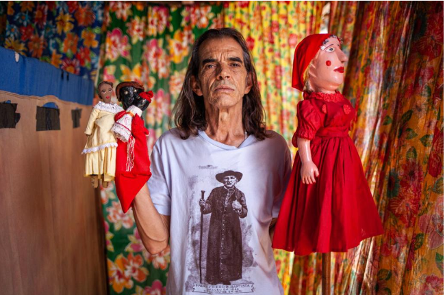 Um homem está em pé com uma expressão séria, segurando bonecos de marionete em ambas as mãos. Ele usa uma camiseta branca com a imagem de uma figura histórica. O fundo é um tecido estampado com cores vibrantes e padrão floral.
