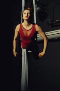 Uma artista de performance está suspensa no ar por tecidos de acrobacia, com um olhar focado para cima. Ela veste um collant vermelho, destacando-se contra um fundo escuro de palco. A iluminação direcionada realça sua expressão intensa e a força física necessária para a pose.