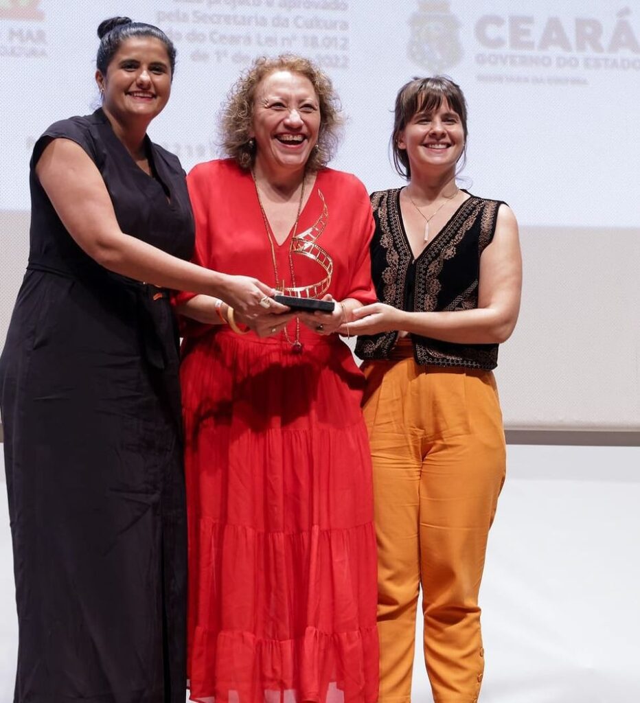 Três mulheres estão no palco com um sorriso radiante, compartilhando um momento de comemoração. A mulher do meio, vestindo um vestido vermelho, segura um troféu com a ajuda de outra mulher à direita, vestida com uma blusa preta sem mangas e calças cor de mostarda. À esquerda, uma terceira mulher em um vestido preto observa a cena com alegria.