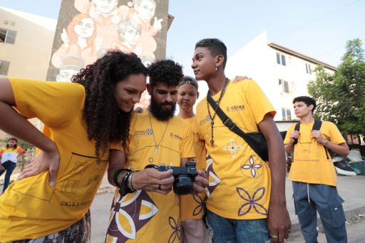 quatro jovens de cammiseta amarela olham uma câmera. Por trás, o prédio de uma cidade com grafite e um jovem de camiseta amarela que olha para o lado.