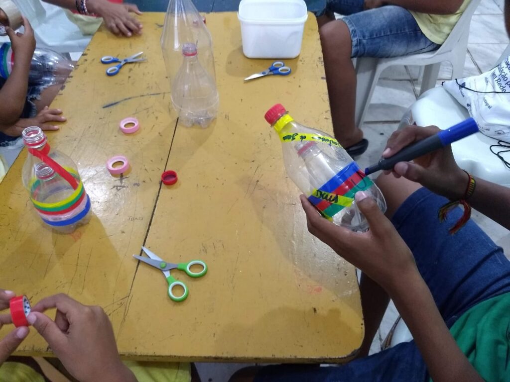 Ao centro da foto, há uma mesa amarela, com garrafas plásticas, tesouras e fitas adesivas coloridas sobre ela. Mãos de crianças aparecem no entorno da mesa produzindo brinquedos com esses materiais.