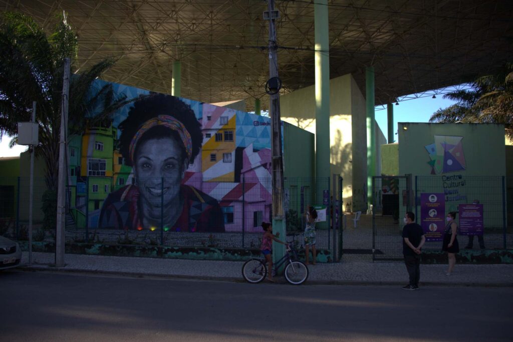Um prédio com fachada verde tem uma das paredes com grafite colorido de Marielle Franco, mulher negra, cabelos crespos e faixa colorida na cabeça. 