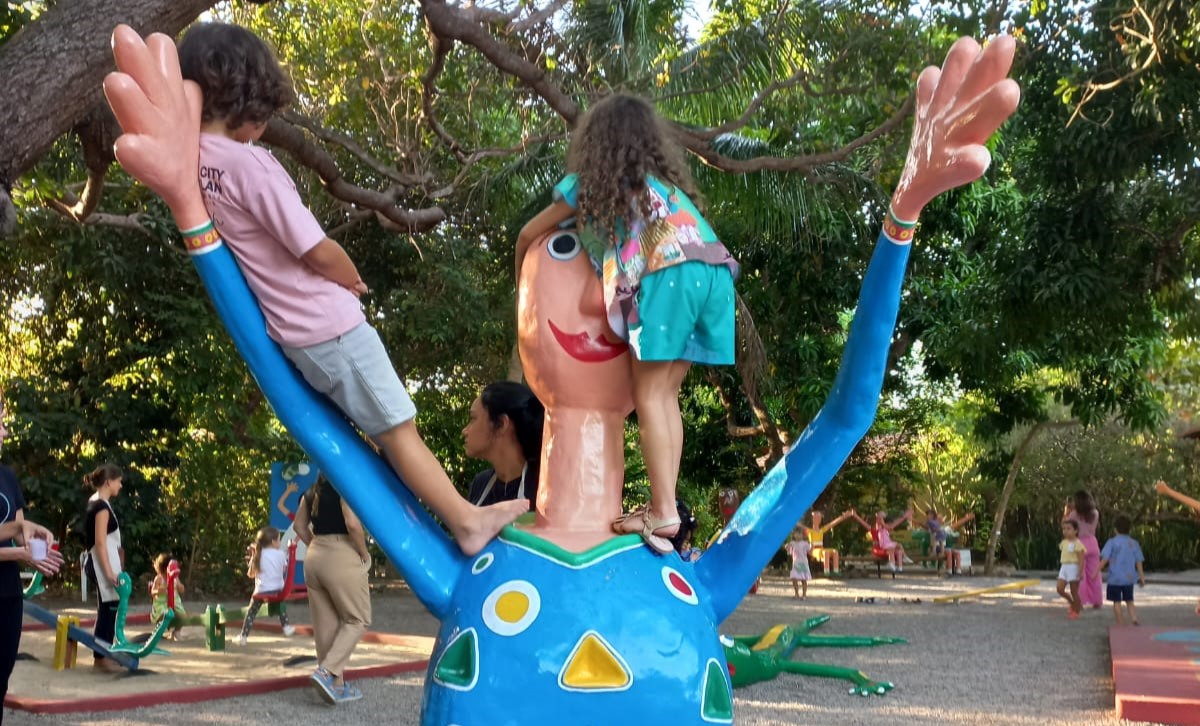 Fotos de crianças no Museu Brinquedim. Duas crianças estão em cima de um brinquedo bem grande. Ele tem o formato de uma pessoa com as mãos erguidas.