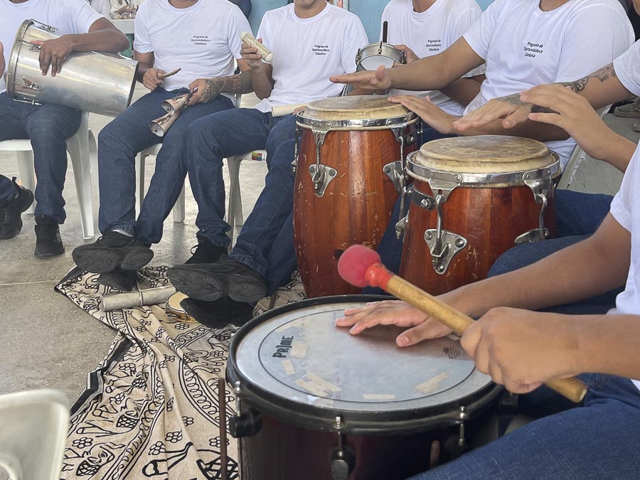 vários jovens do sexo masculino tocam instrumentos de percussão, estão de camisa branca e somente suas mãoes e tórax aparecem na foto.