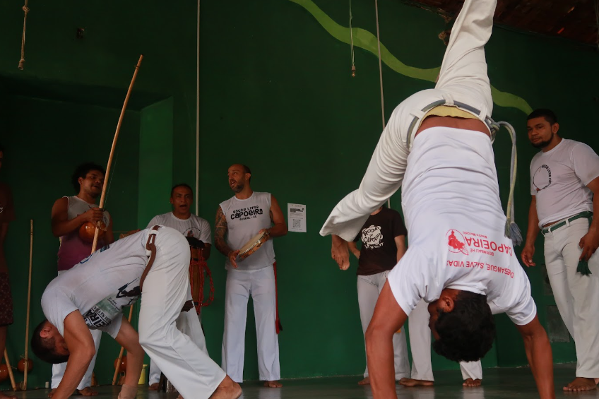 Foto de roda de capoeira. Dois rapazes estão no centro fazendo movimentos típicos da prática. Todos usam roupas brancas. No fundo três pessoas tocam beribal.