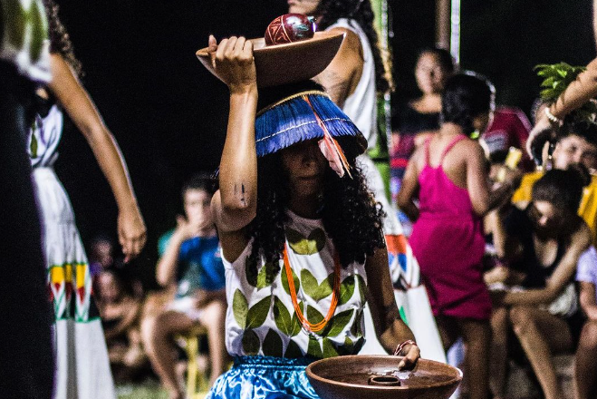 Imagem do Ponto de Cultura Recanto dos Encantados, espaço apoiado pela Temporada de Arte Cearense 2023/24. Uma mulher segura uma cumbuca na cabeça. Ela está com adorno azul na cabeça.