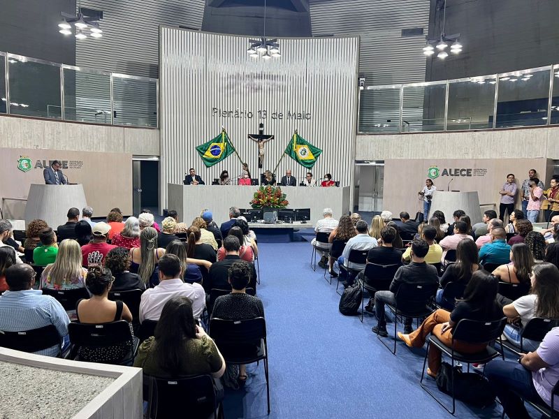 A imagem mostra uma cerimônia em um auditório com cadeiras ocupadas por espectadores voltados para a frente. Há uma grande mesa ao fundo com pessoas sentadas, e um homem está de pé falando ao microfone. O local é amplo e decorado com as bandeiras do Brasil e do estado do Ceará, além do logo da Assembleia Legislativa do Estado do Ceará (ALCE). Há pessoas em pé nas laterais observando o evento.