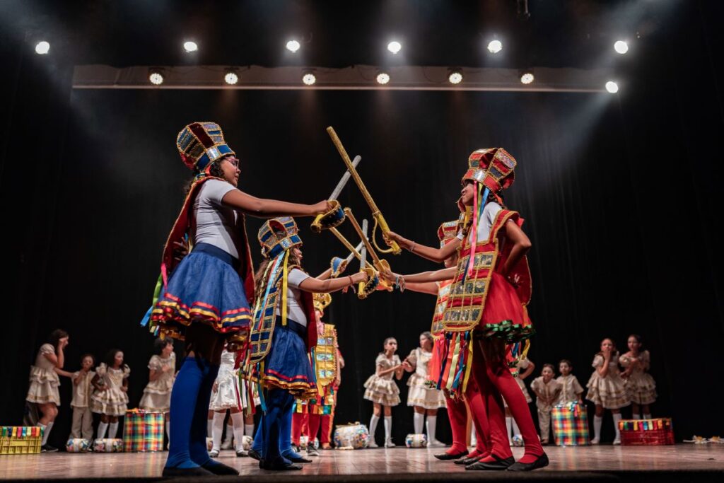 Na imagem, crianças vestidas com trajes coloridos estão realizando uma apresentação de dança. No centro, quatro crianças em roupas adornadas com fitas e tecidos brilhantes seguram bastões que se encontram no meio, criando uma formação em "X". Ao fundo, outras crianças em uniforme escolar observam e aplaudem. A iluminação do palco foca nos dançarinos, destacando a vivacidade das cores e o movimento da dança.