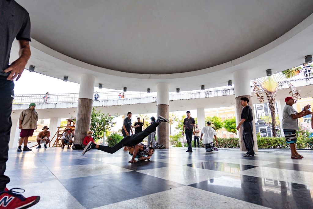 A fotografia captura um grupo de pessoas dançando breakdance em uma área coberta por uma estrutura circular com colunas. No centro, uma pessoa está realizando um movimento de dança no ar, apoiando-se apenas em uma mão, enquanto os outros observam ou esperam sua vez. A cena transmite energia e movimento e parece ser uma prática informal ou encontro social de dançarinos. Ao fundo, podemos ver um ambiente urbano com palmeiras e edifícios.